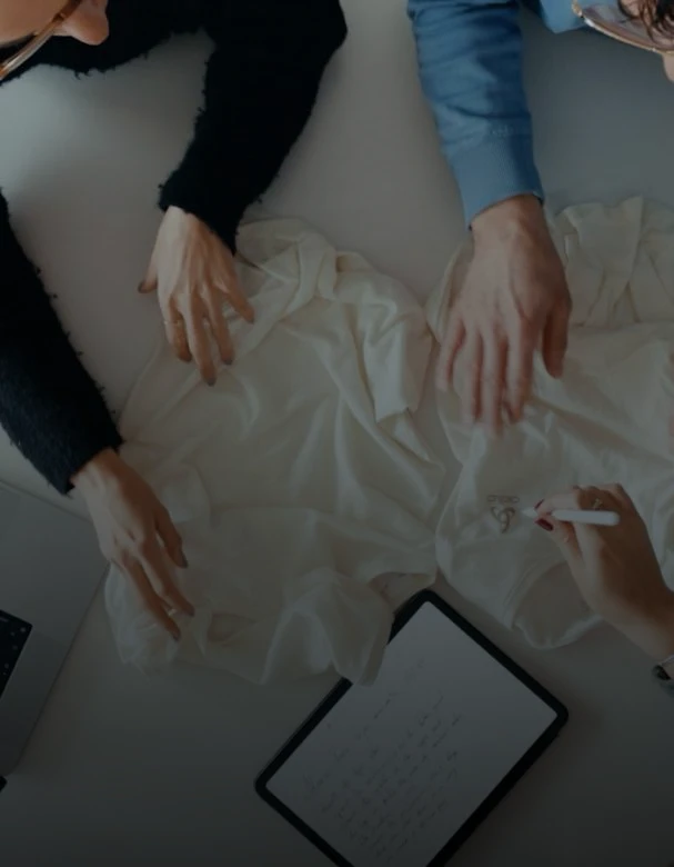 Bovenaanzicht van twee mensen die witte shirts op een bureau bekijken. Een van hen wijst met een stylus naar een logo naast een tablet met notities.