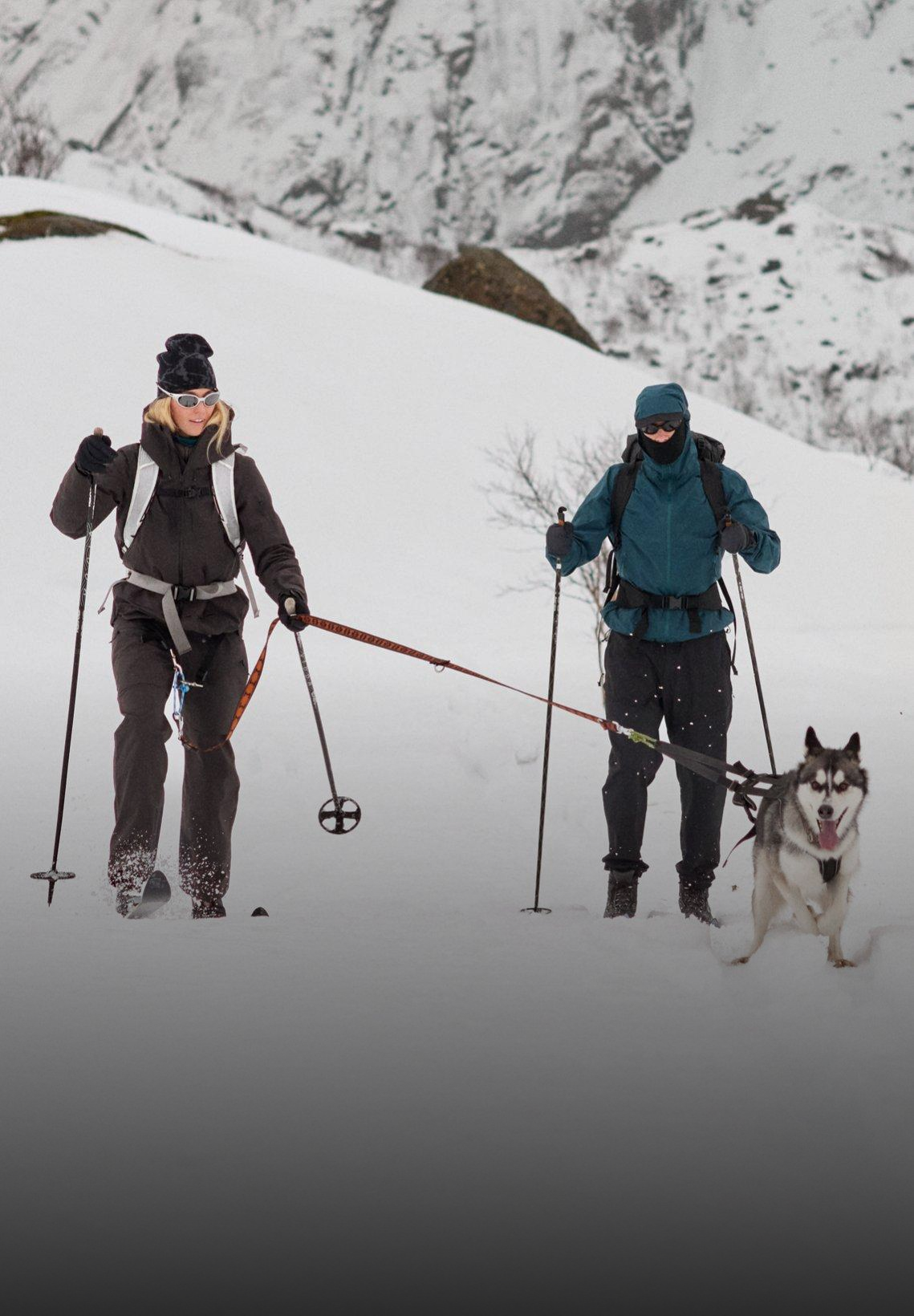 Two people skiing in a snowy mountain landscape. A husky on a lead pulls a woman in black, alongside a person in blue.