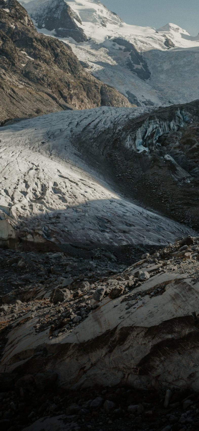 A hiker stands on a rocky outcrop, gazing at a sweeping glacier and towering snow-capped mountains in a rugged landscape.