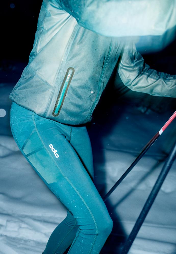 Une personne fait du ski de fond de nuit dans la neige, vêtue d'une veste imperméable Dual Dry et d'un legging Odlo assorti de couleur sarcelle, tenant des bâtons de ski.