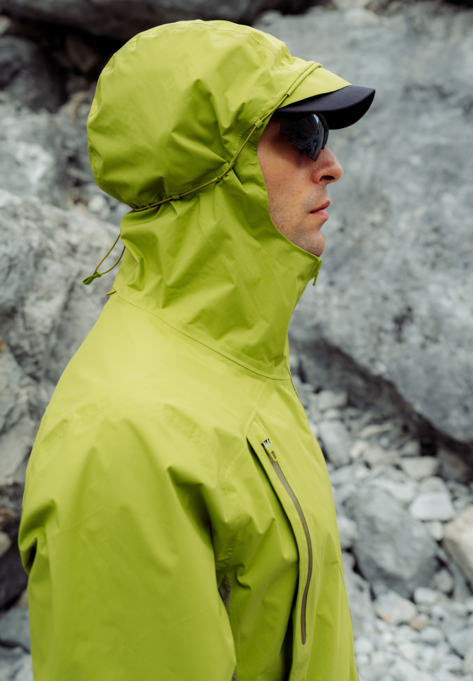 Profile of a man wearing a bright green hooded running jacket, black cap, and sunglasses, standing against grey rocks.
