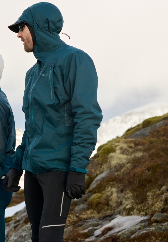 Man in a teal hooded jacket, cap, and black tights stands on a rocky, mossy hill with snowy mountains behind him.