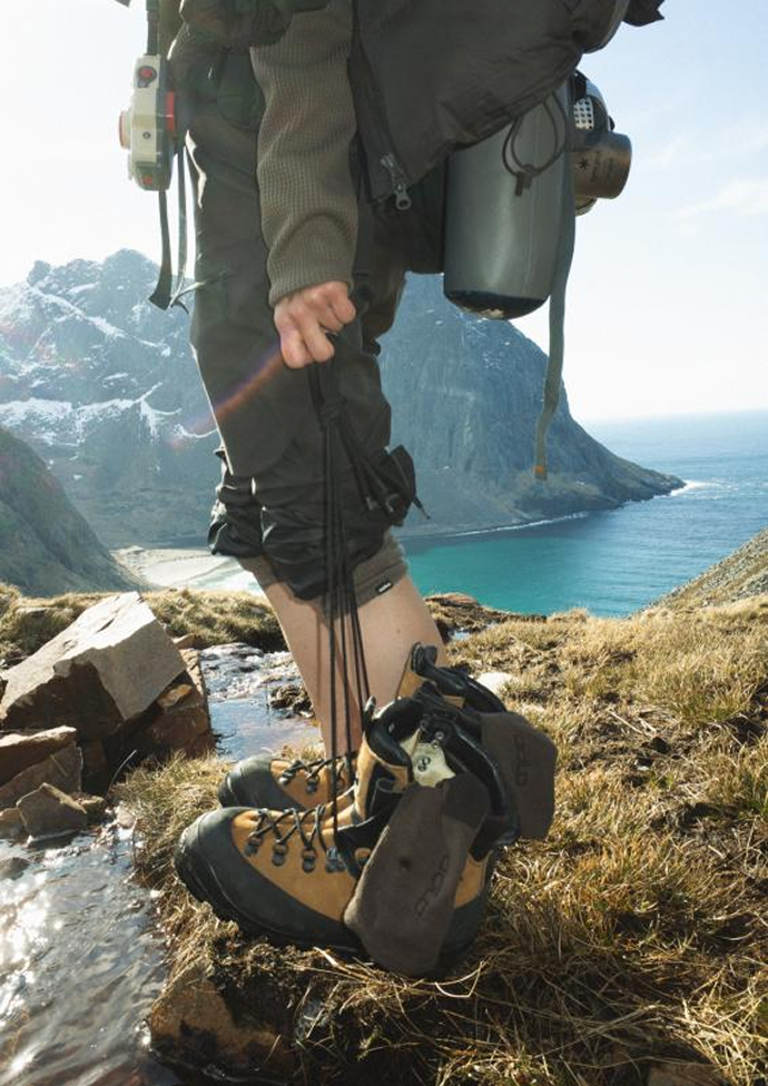 Barefoot hiker with a backpack holds brown boots by a stream, overlooking a blue bay and snowy mountains.