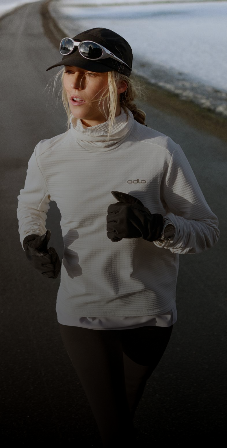 Une femme qui court sur une route bordée de neige, portant un haut blanc à manches longues, une casquette noire, des lunettes de soleil et des gants noirs.