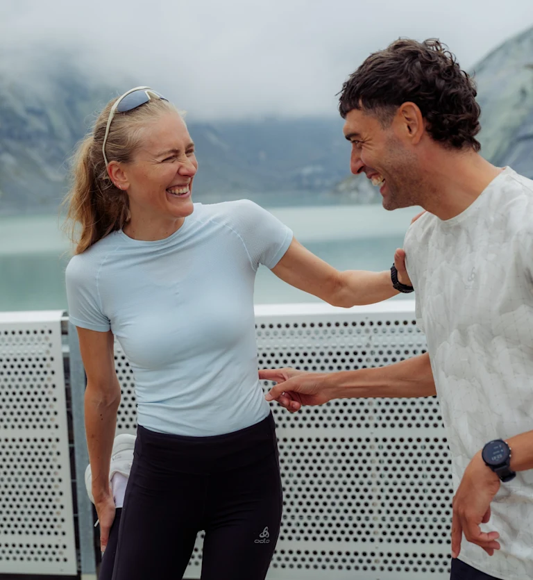 A blonde woman in a light blue t-shirt and a man in a white t-shirt laugh playfully outdoors near a mountain lake.
