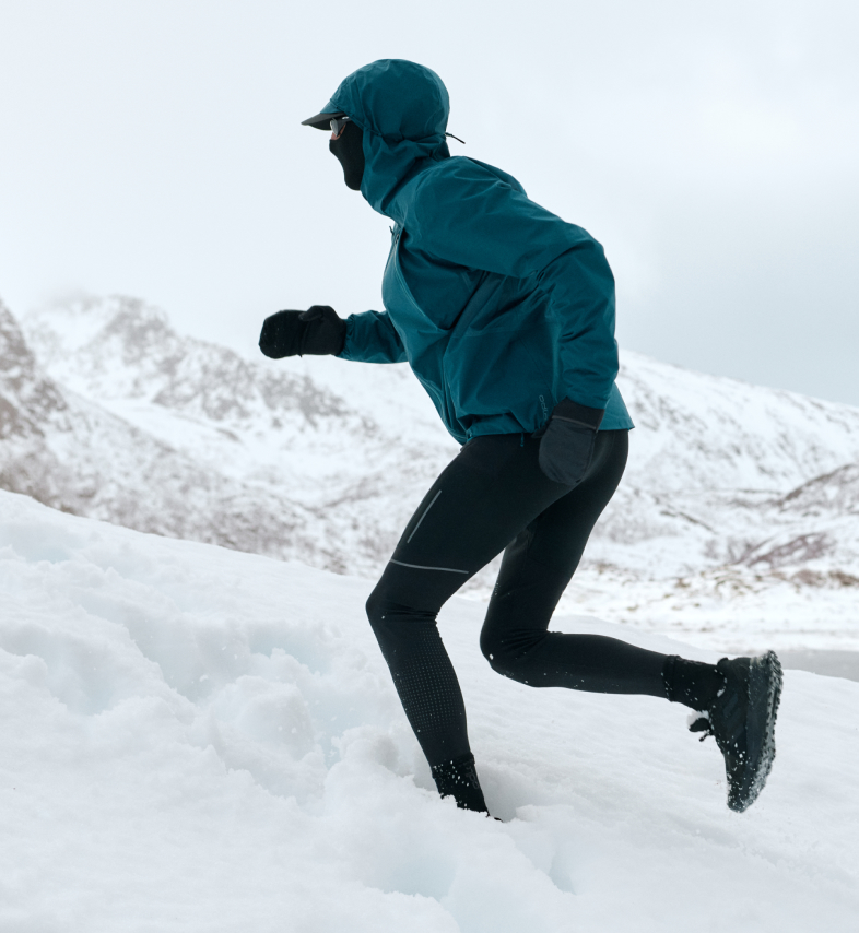 Person in einer türkisfarbenen Kapuzenjacke und schwarzen Leggings läuft bergauf durch tiefen Schnee in einer winterlichen Berglandschaft.