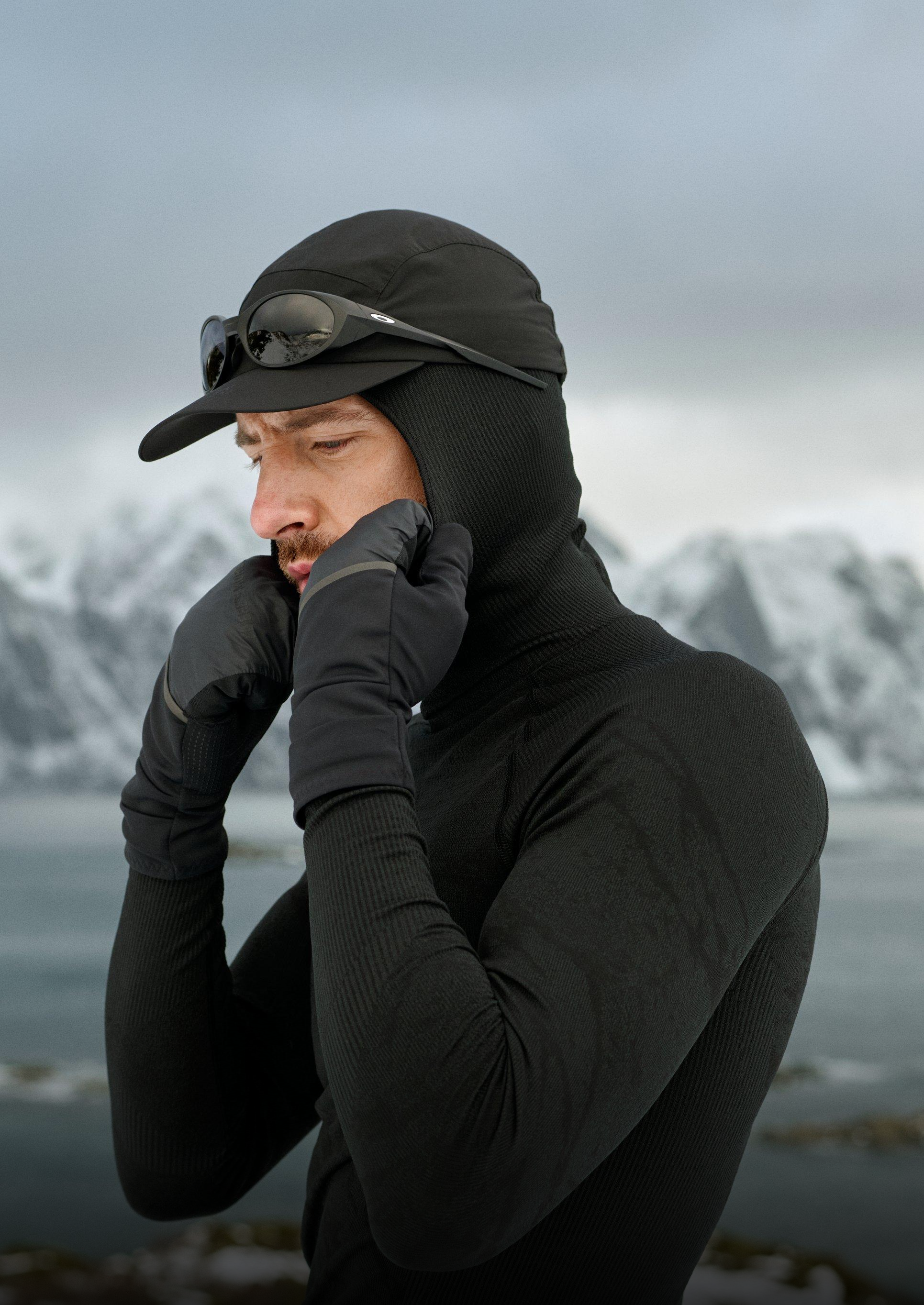Mann in schwarzem Blackcomb Base Layer, Cap und Handschuhen, der seine Gesichtsmaske richtet, mit verschneiten Bergen und Wasser im Hintergrund.