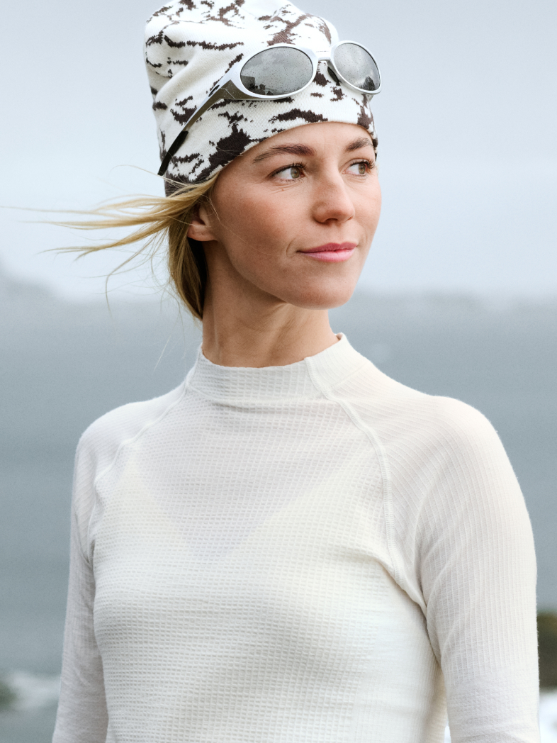 Blonde woman in a white base layer and patterned beanie with sunglasses. Wind blows her hair against a grey background.