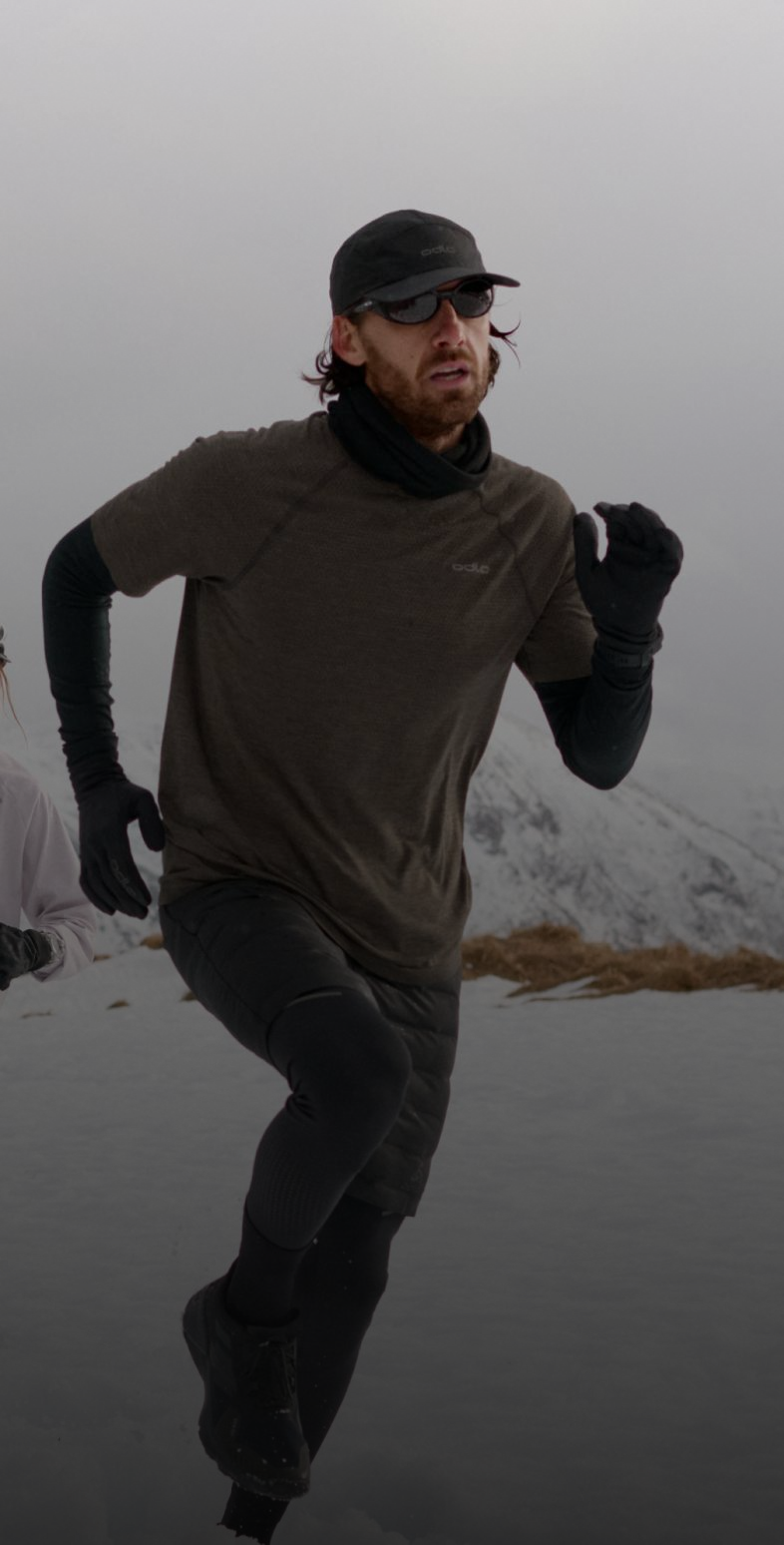 Two runners in winter gear sprint through deep snow, the man leading. Snowy mountains and cloudy sky form a rugged, cold backdrop.