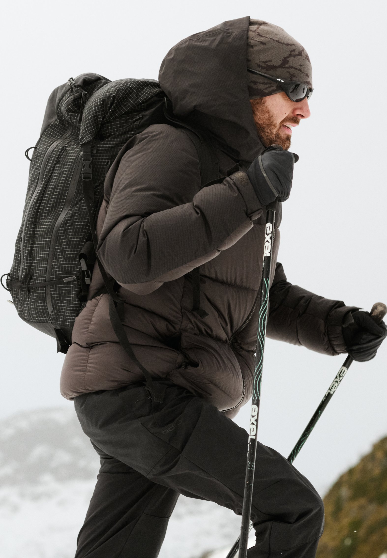 Ein Mann in dunkler Winterbekleidung und eine Frau in einer türkisfarbenen Jacke, beide mit Beanies und Rucksäcken unter einem grauen Himmel.
