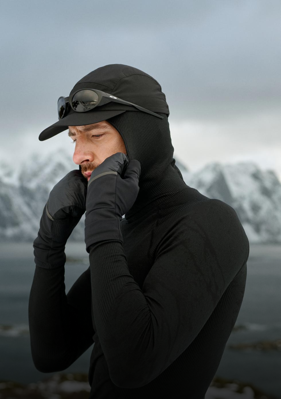 Mann in schwarzem Blackcomb Base Layer, Handschuhen und Mütze richtet seine Kapuze vor dem Hintergrund eines kalten Meeres und schneebedeckter Berge.