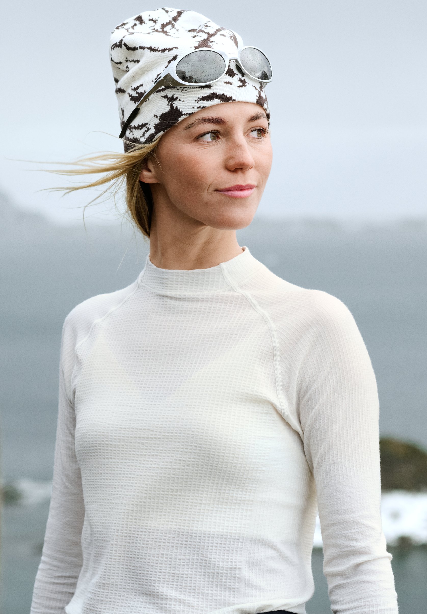 Frau in einem weißen Thermo-Shirt und gemusterter Beanie, mit Sonnenbrille auf dem Kopf, blickt nach rechts, Haare wehen im Wind.