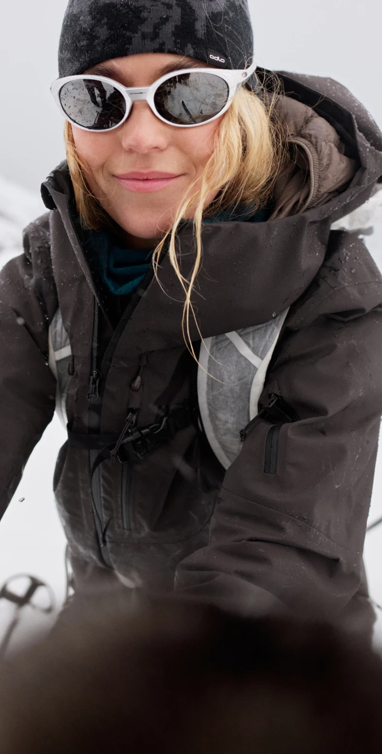 Primo piano di una donna bionda sorridente nella neve, con un berretto nero, occhiali da sole bianchi e un giaccone invernale scuro.