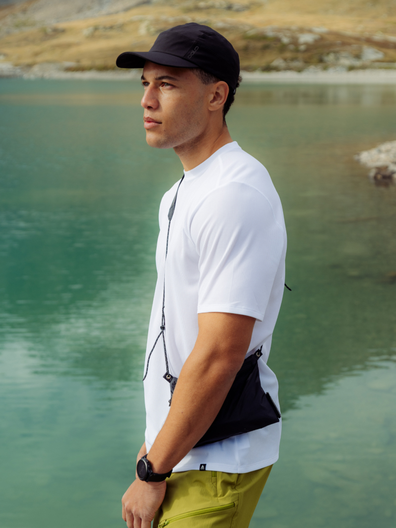Man in a white and light base layer t-shirt, olive shorts, and black cap stands by a green mountain lake, wearing a small black crossbody bag.
