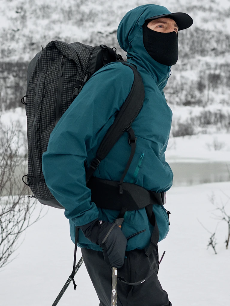 Two people in winter gear against a grey sky. A woman in a teal jacket stands in front of a bearded man in a dark jacket.