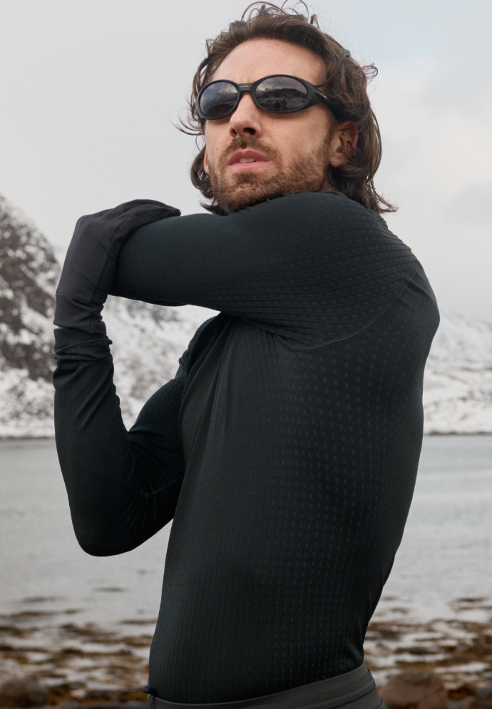 A bearded man in black athletic wear and sunglasses stretches his arm outdoors near water and snow-covered mountains.