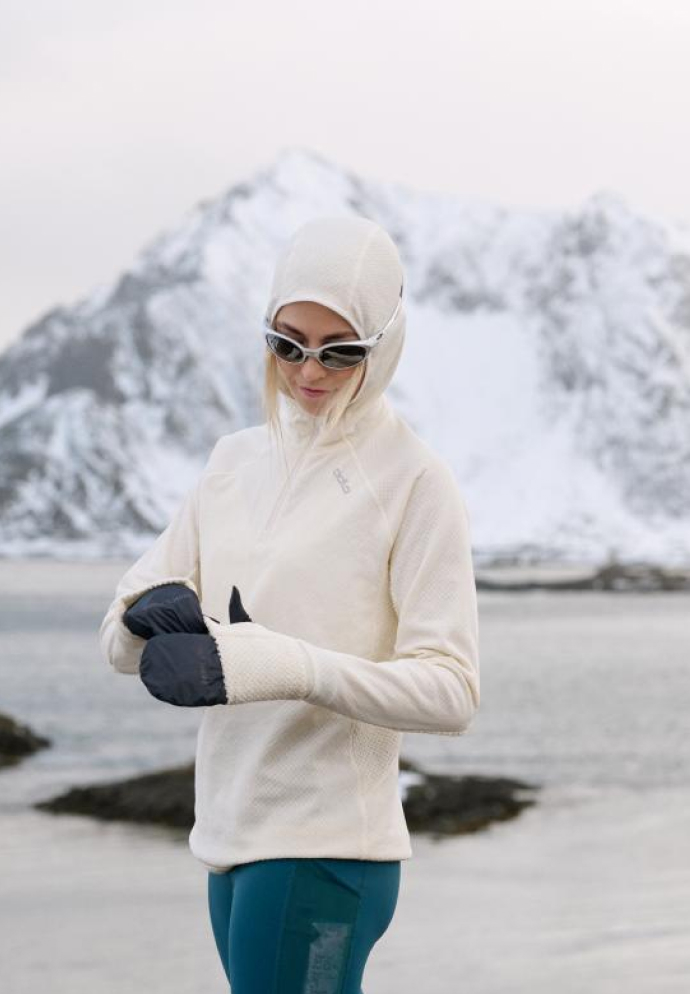 Une femme en polaire blanche à capuche, lunettes de soleil et leggings sarcelle ajuste ses moufles foncées près de l'eau, avec des montagnes enneigées en arrière-plan.