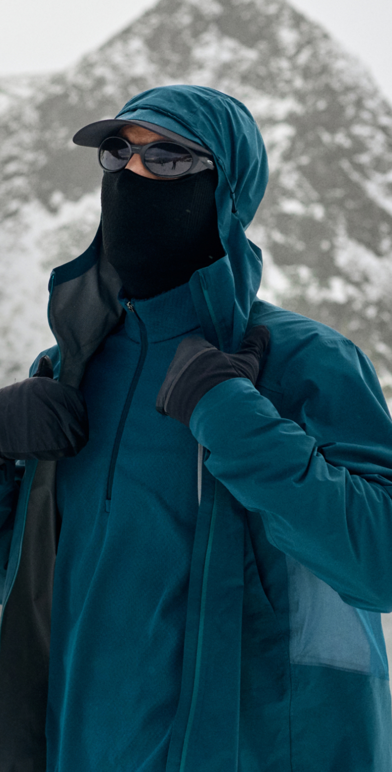 Person im Schnee, trägt eine türkise Kapuzenjacke, eine schwarze Sonnenbrille, eine Gesichtsmaske und Handschuhe, richtet den Kragen zurecht.