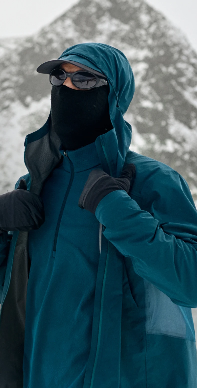 Person im Schnee, trägt eine türkise Kapuzenjacke, eine schwarze Sonnenbrille, eine Gesichtsmaske und Handschuhe, richtet den Kragen zurecht.