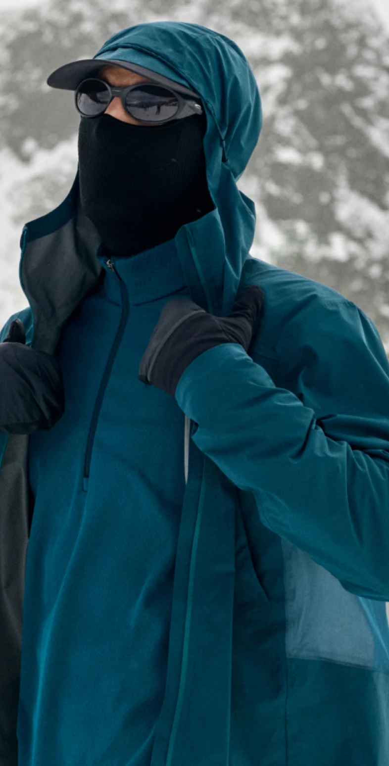 Persona sulla neve che indossa una giacca con cappuccio color verde acqua, occhiali da sole neri, una maschera per il viso e guanti, mentre sistema il colletto.