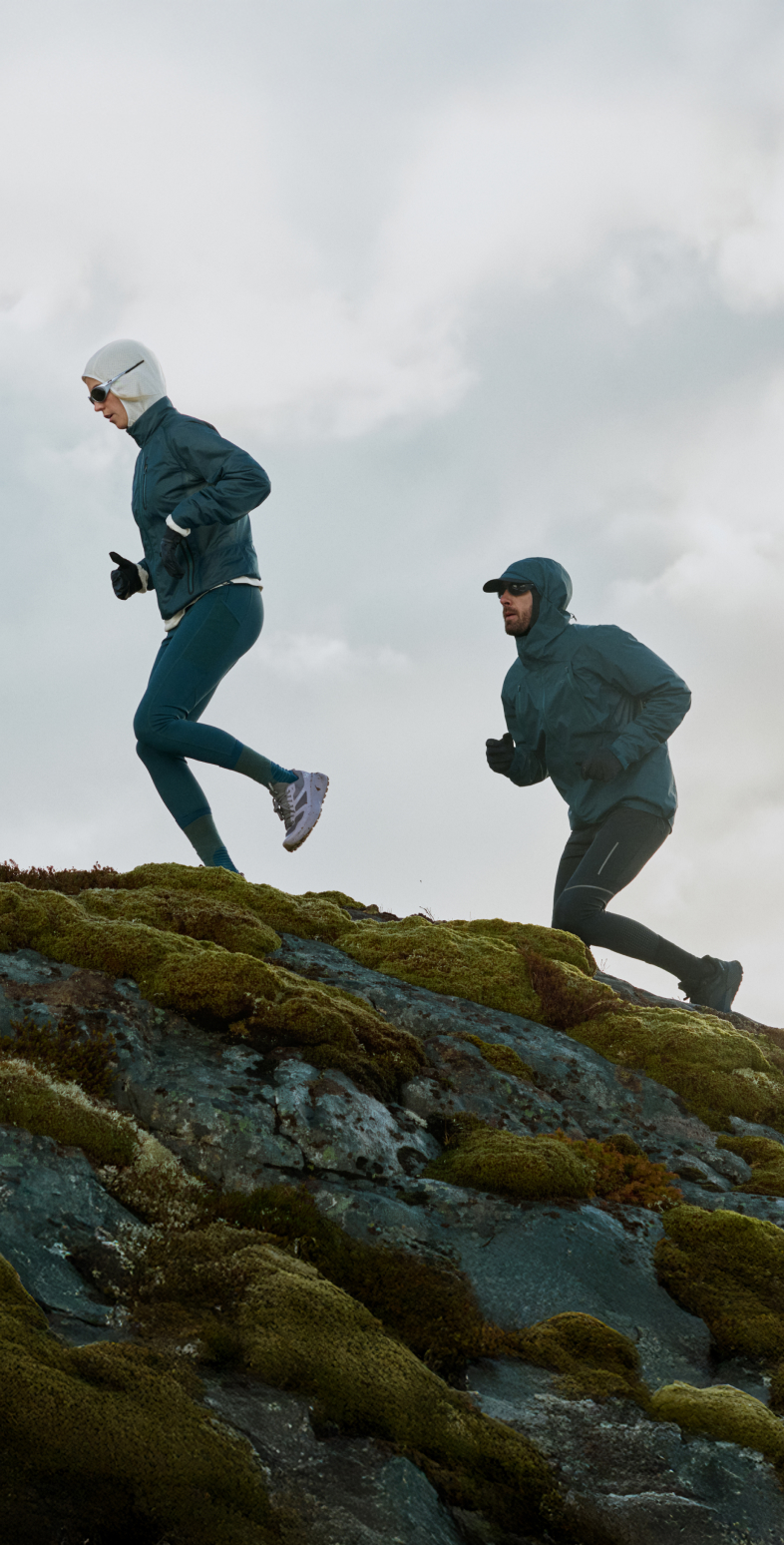 Two people trail running uphill on mossy rocks under a cloudy sky, wearing dark blue jackets and cold-weather gear.