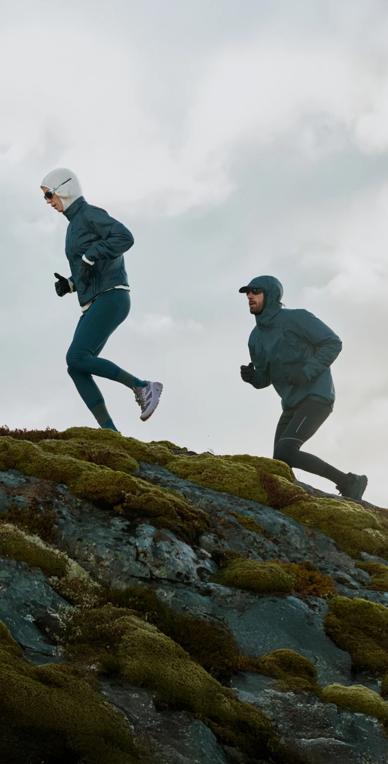 Two people trail running uphill on mossy rocks under a cloudy sky, wearing dark blue jackets and cold-weather gear.
