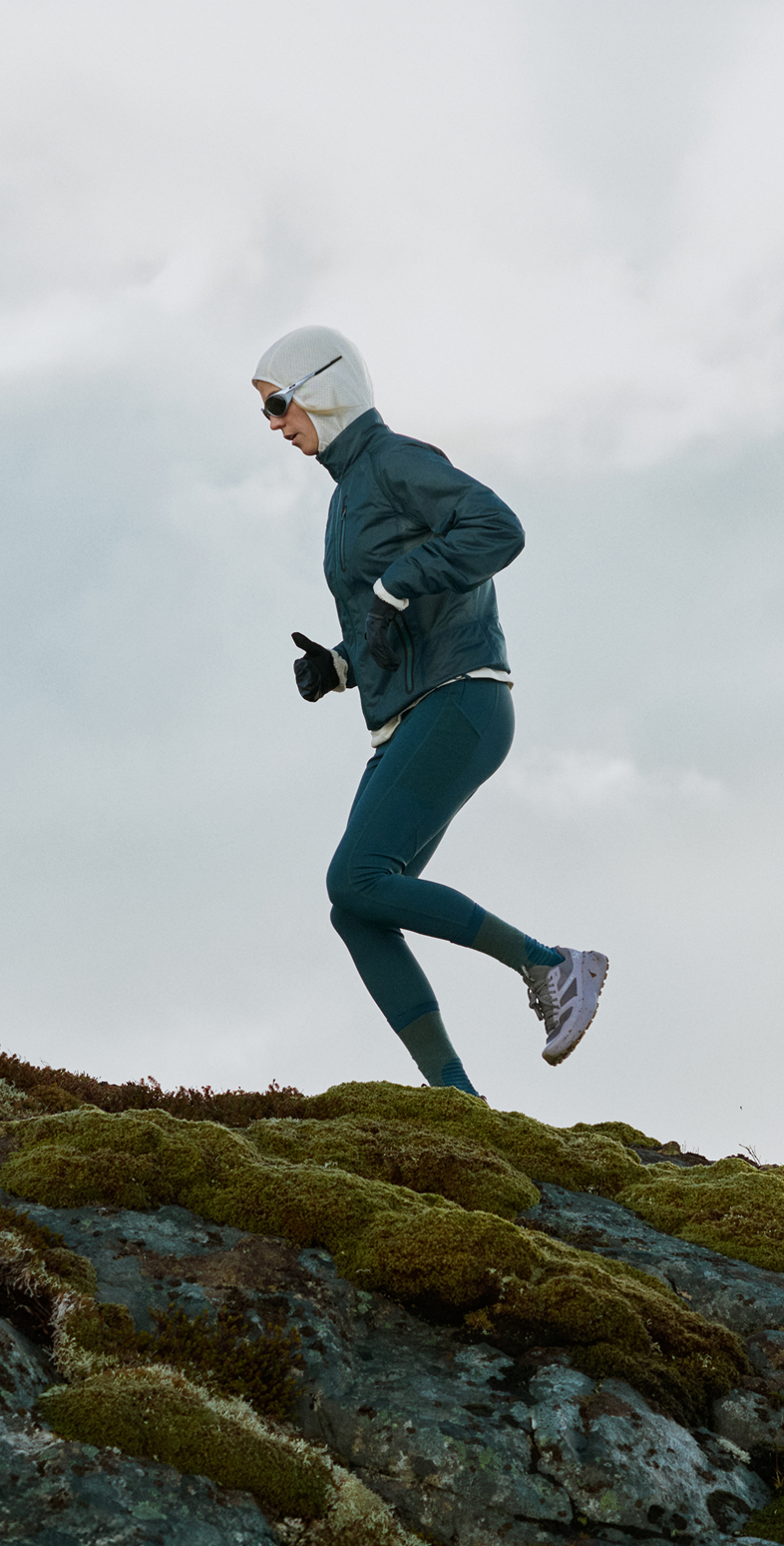 Zwei Personen beim Trailrunning bergauf über bemooste Felsen unter bewölktem Himmel, in dunkelblauen Jacken und Kälteschutzbekleidung.