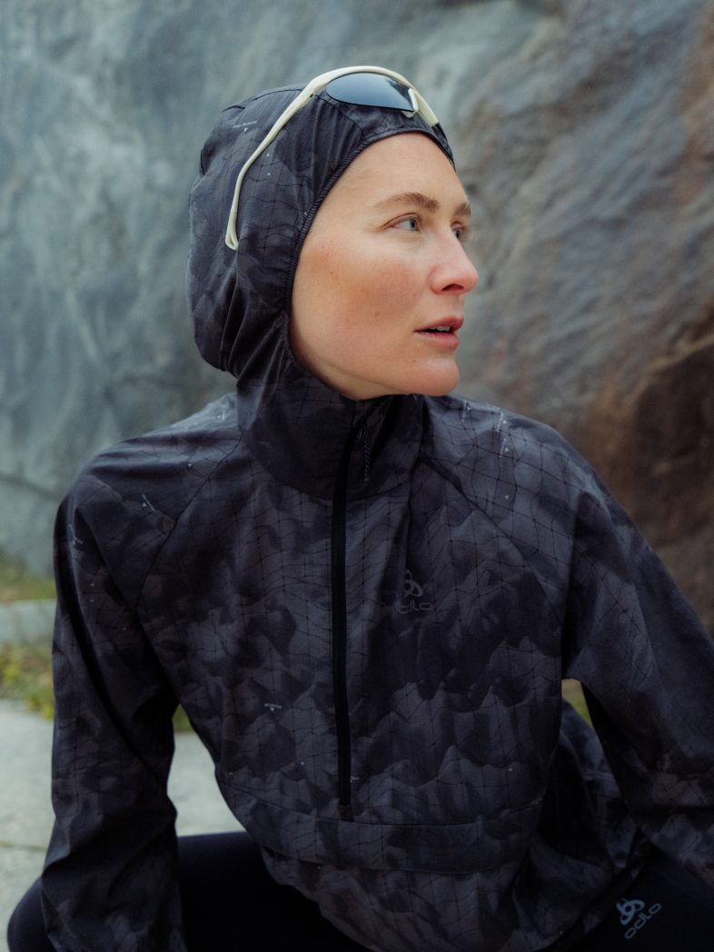 Woman in a dark patterned hooded running jacket with sunglasses on the hood looks to the side against a rocky background.