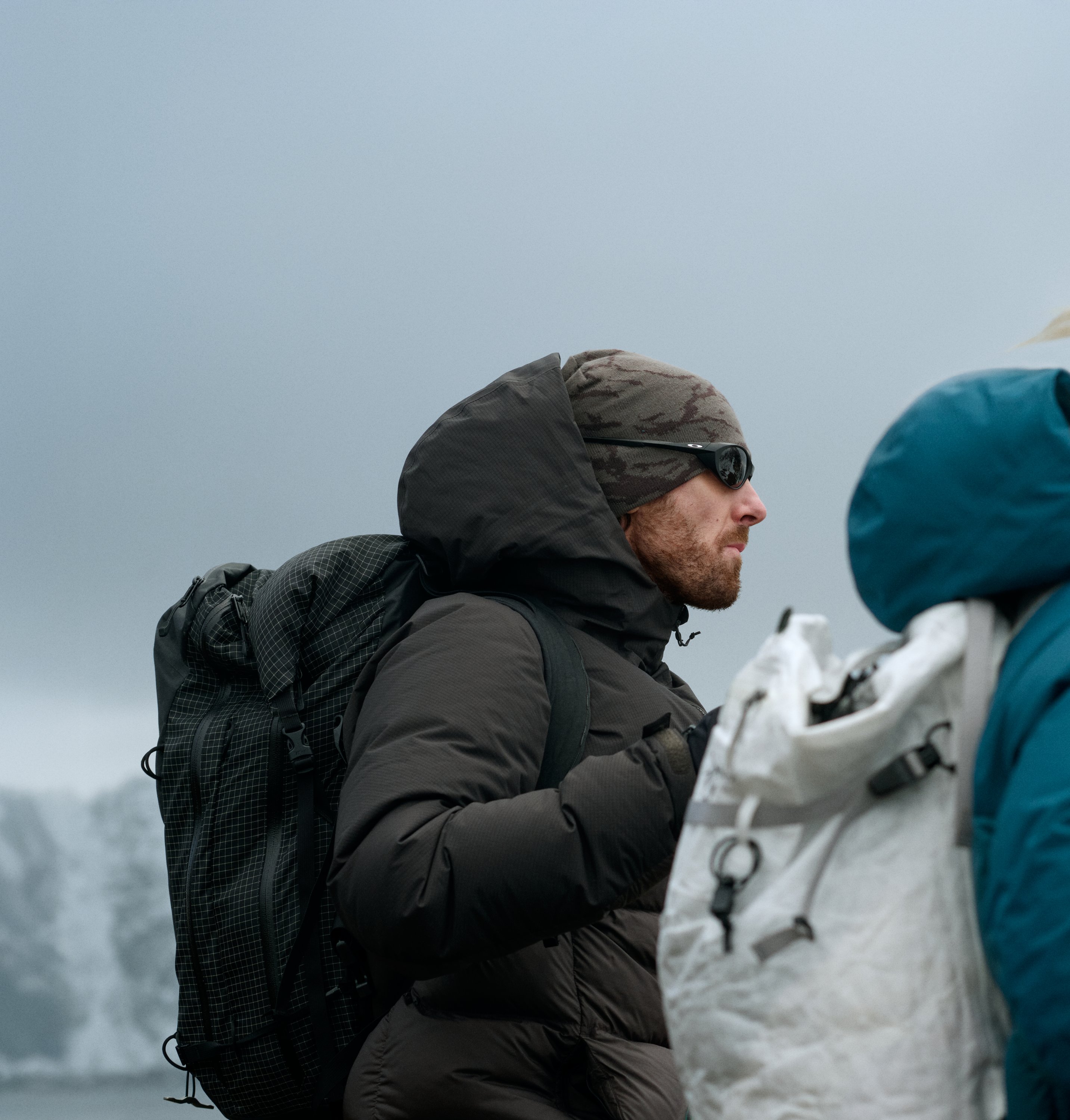 A man in a dark jacket and a woman in a teal jacket, both wearing beanies and backpacks, look right against a grey sky.
