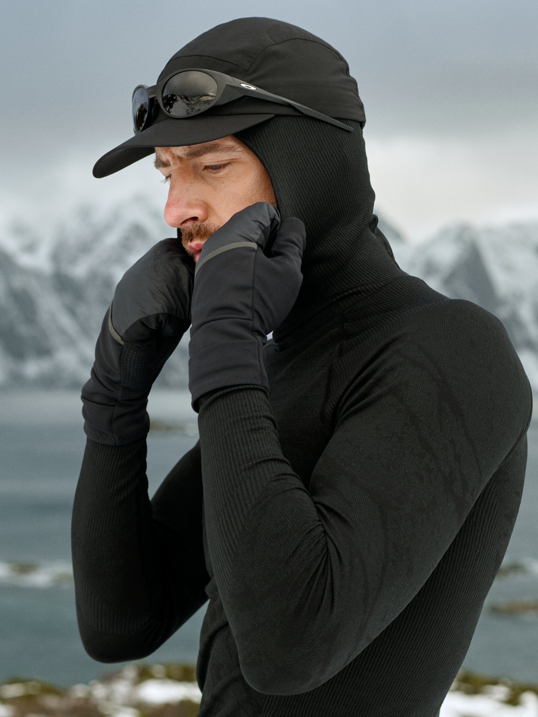 Man in zwarte performancekleding en met zonnebril rekt zijn arm uit op een rotsachtige oever, met water en besneeuwde bergen op de achtergrond.