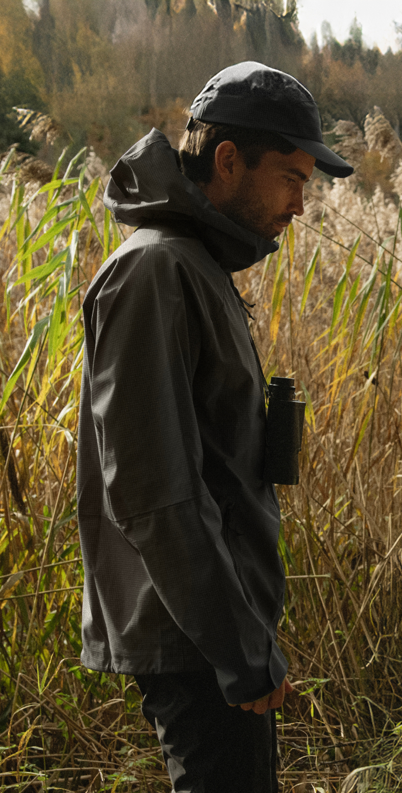 Profil eines Mannes in einer dunkelgrauen Jacke und Mütze, der im hohen Schilf steht und ein Fernglas um den Hals trägt.