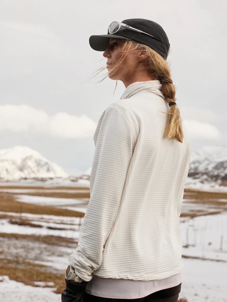 Woman in a white fleece mid layer and black cap with goggles stands outdoors, looking sideways over a snowy, mountainous landscape under cloudy sky.