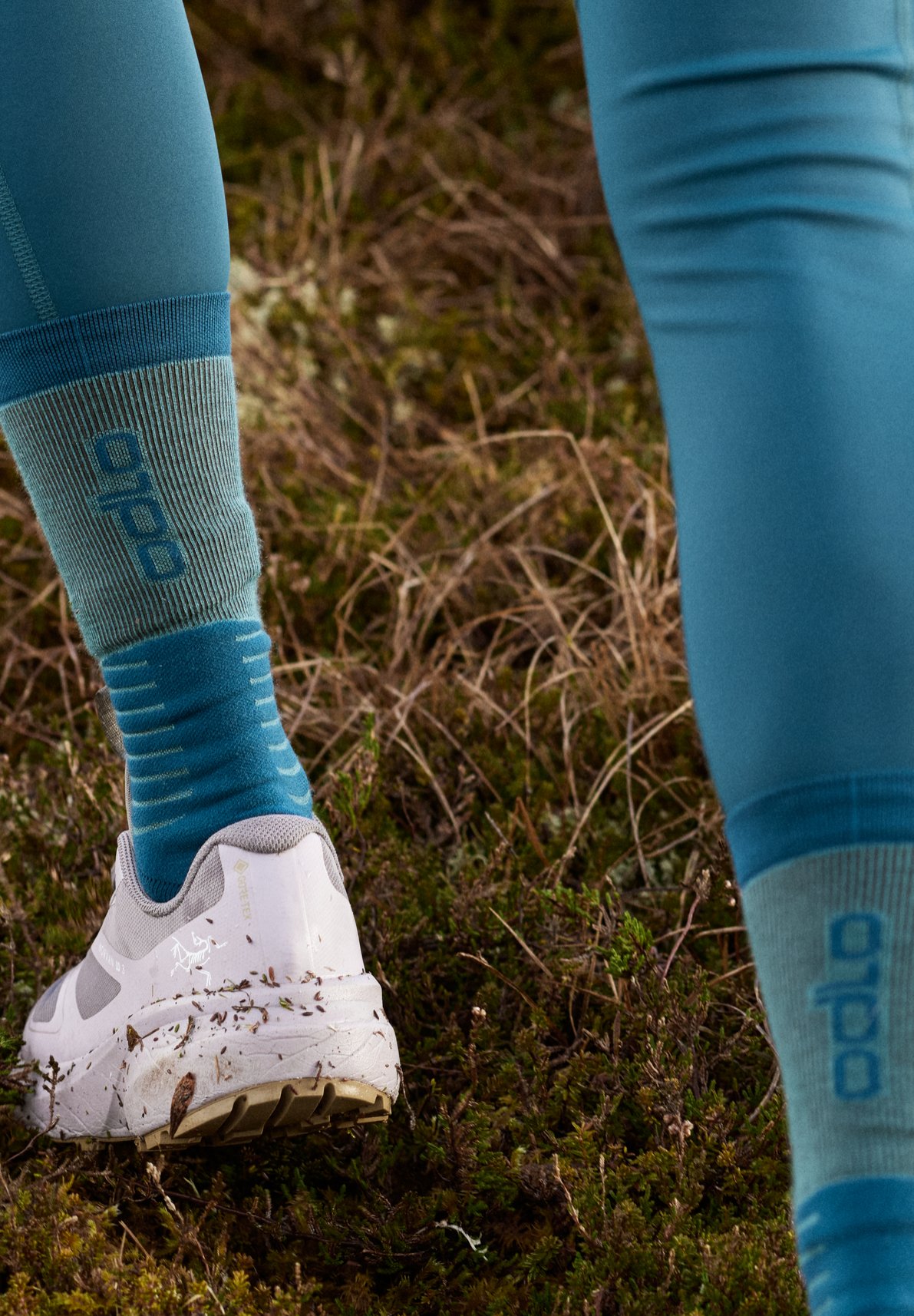 Benen in een petrolblauwe legging, blauwe Odlo-sokken en modderige witte trailrunningschoenen op grasachtig terrein.