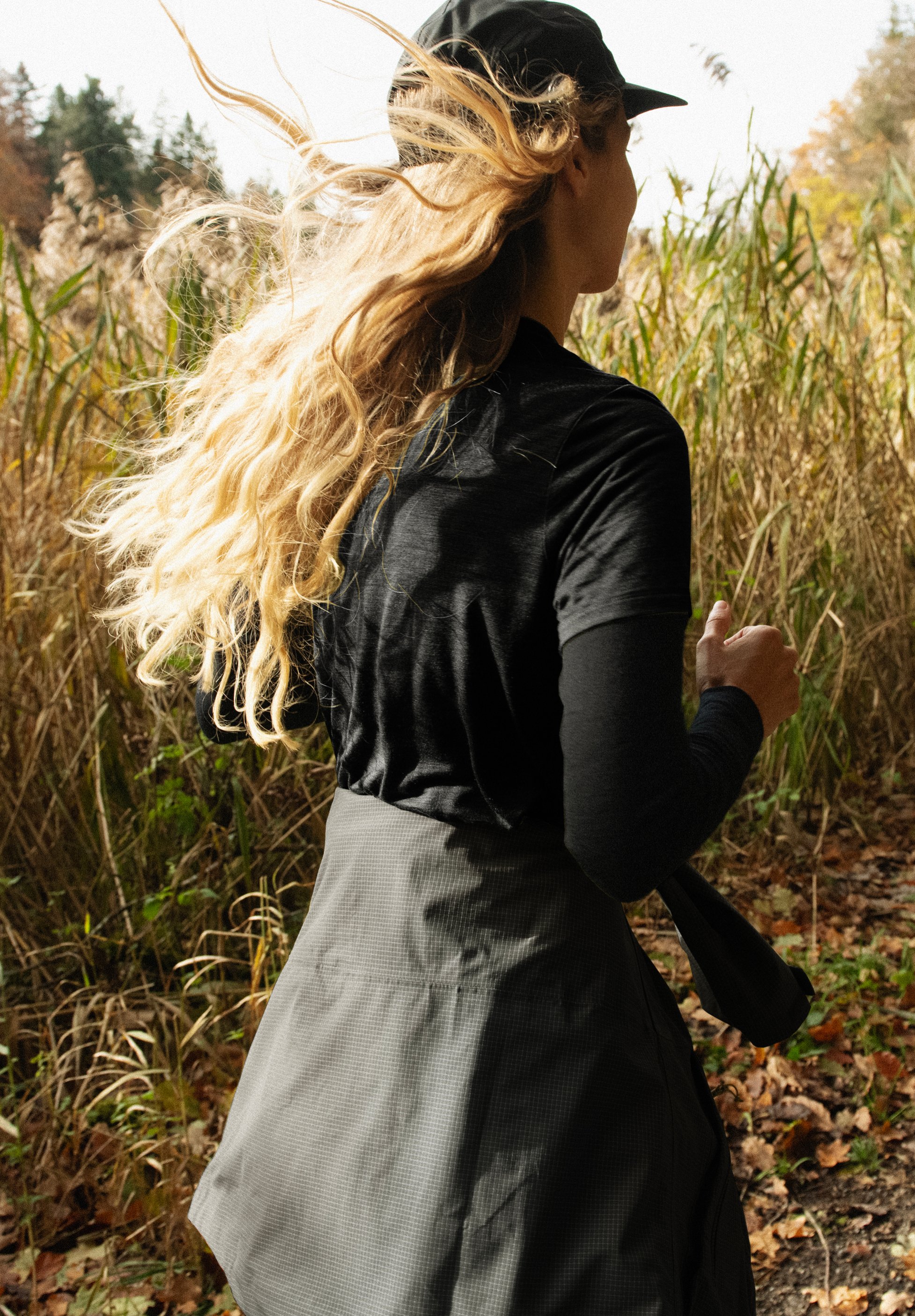 Woman running on an autumn trail through tall grass. She wears a black cap and shirt, her long blonde hair blowing.