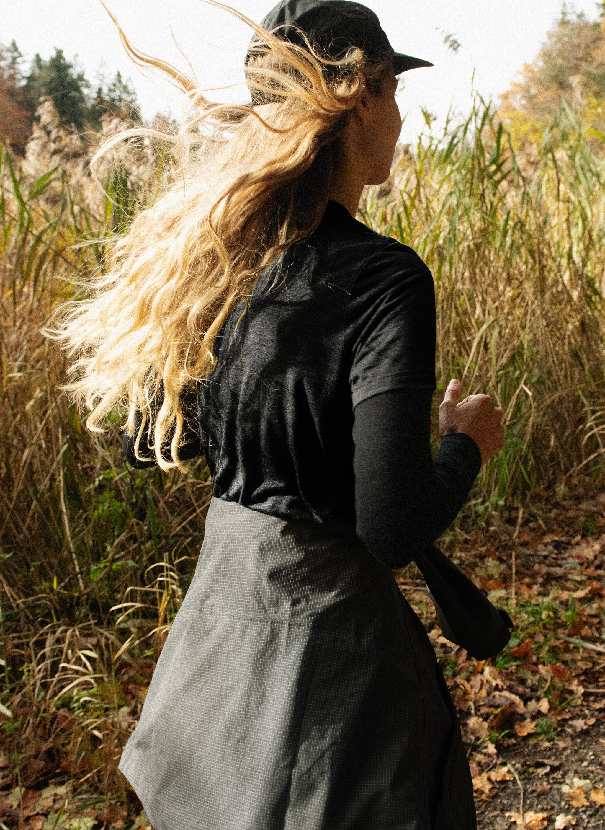 Donna che corre su un sentiero autunnale tra l'erba alta. Indossa un berretto e una maglietta neri, i lunghi capelli biondi al vento.