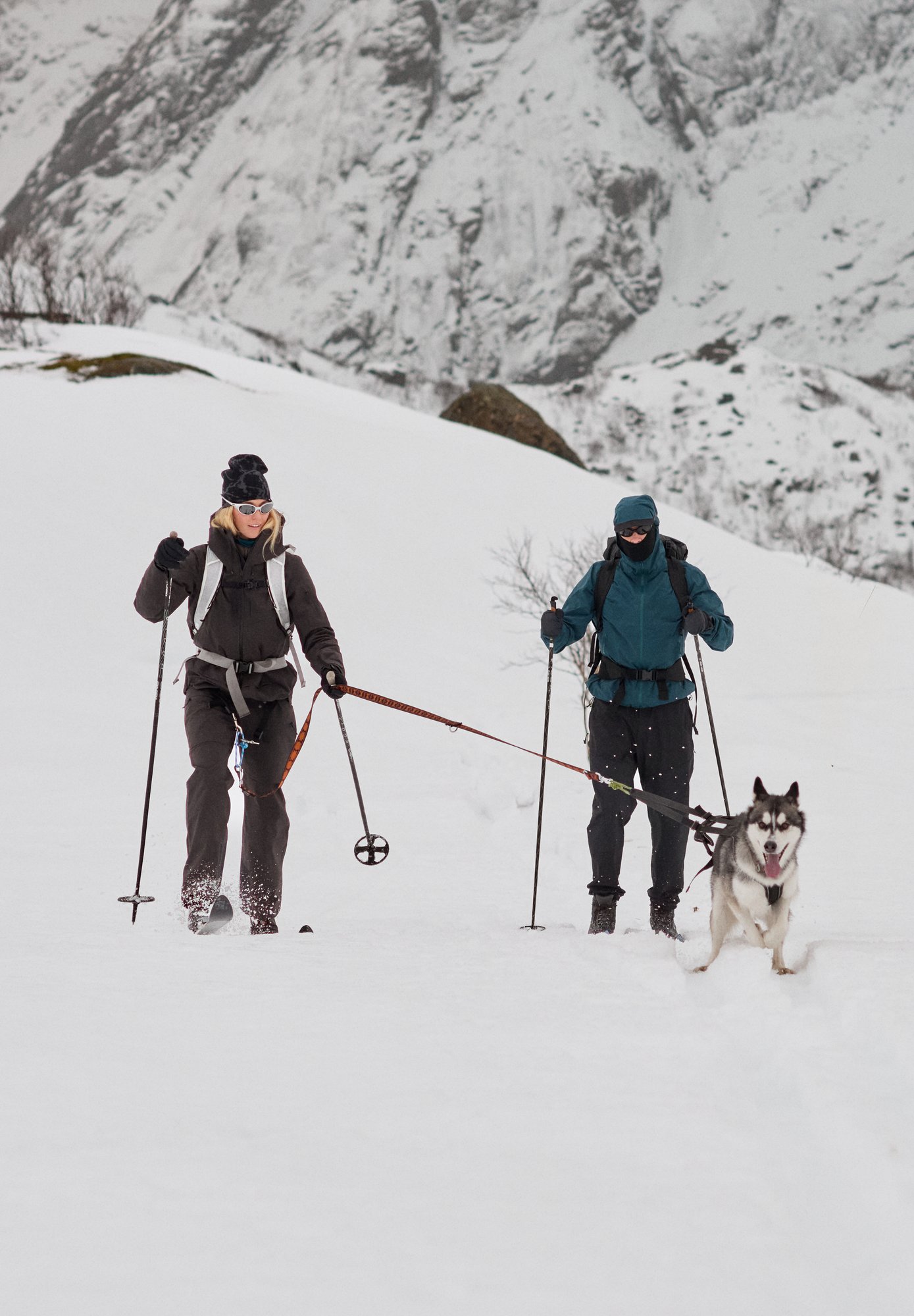 Eine Frau in Schwarz und eine Person in Blau fahren in einer verschneiten Berglandschaft Ski, gezogen von einem Husky an einem Geschirr.