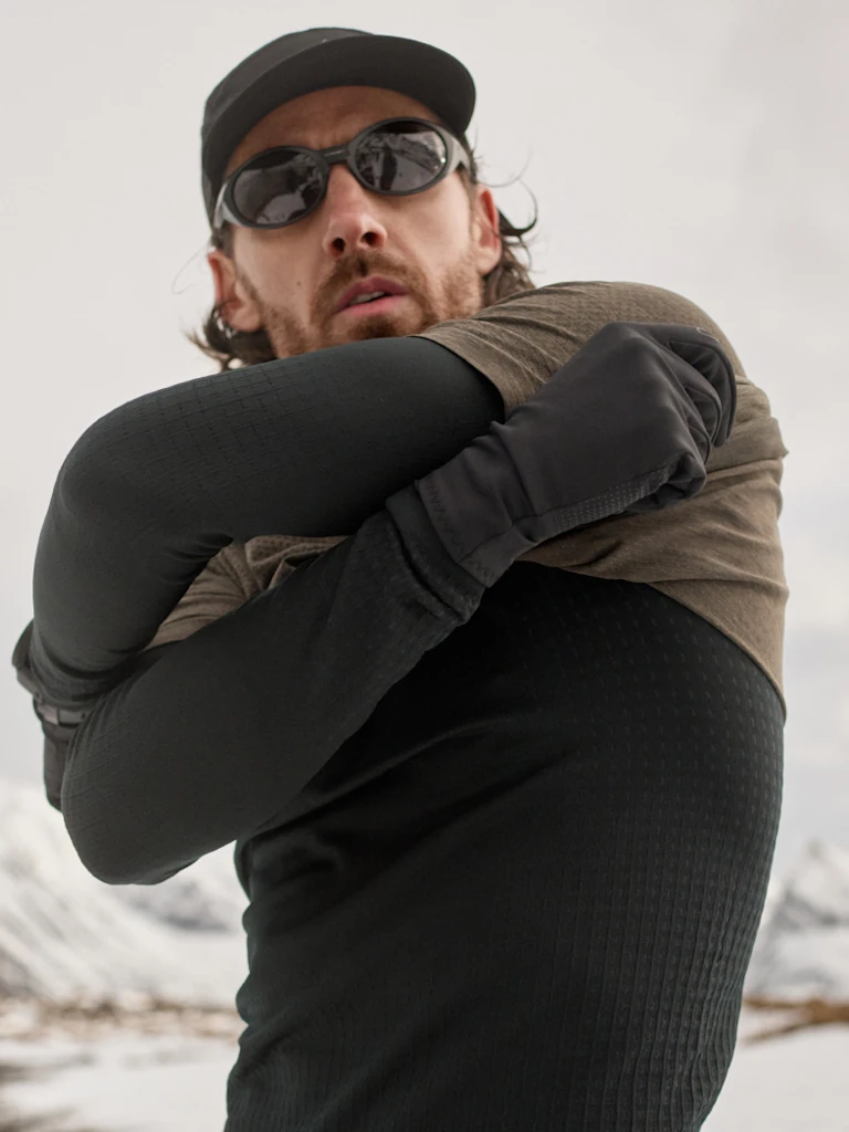Bearded man in cap, sunglasses and gloves pulls an olive t-shirt over a black base layer in a snowy mountain landscape.
