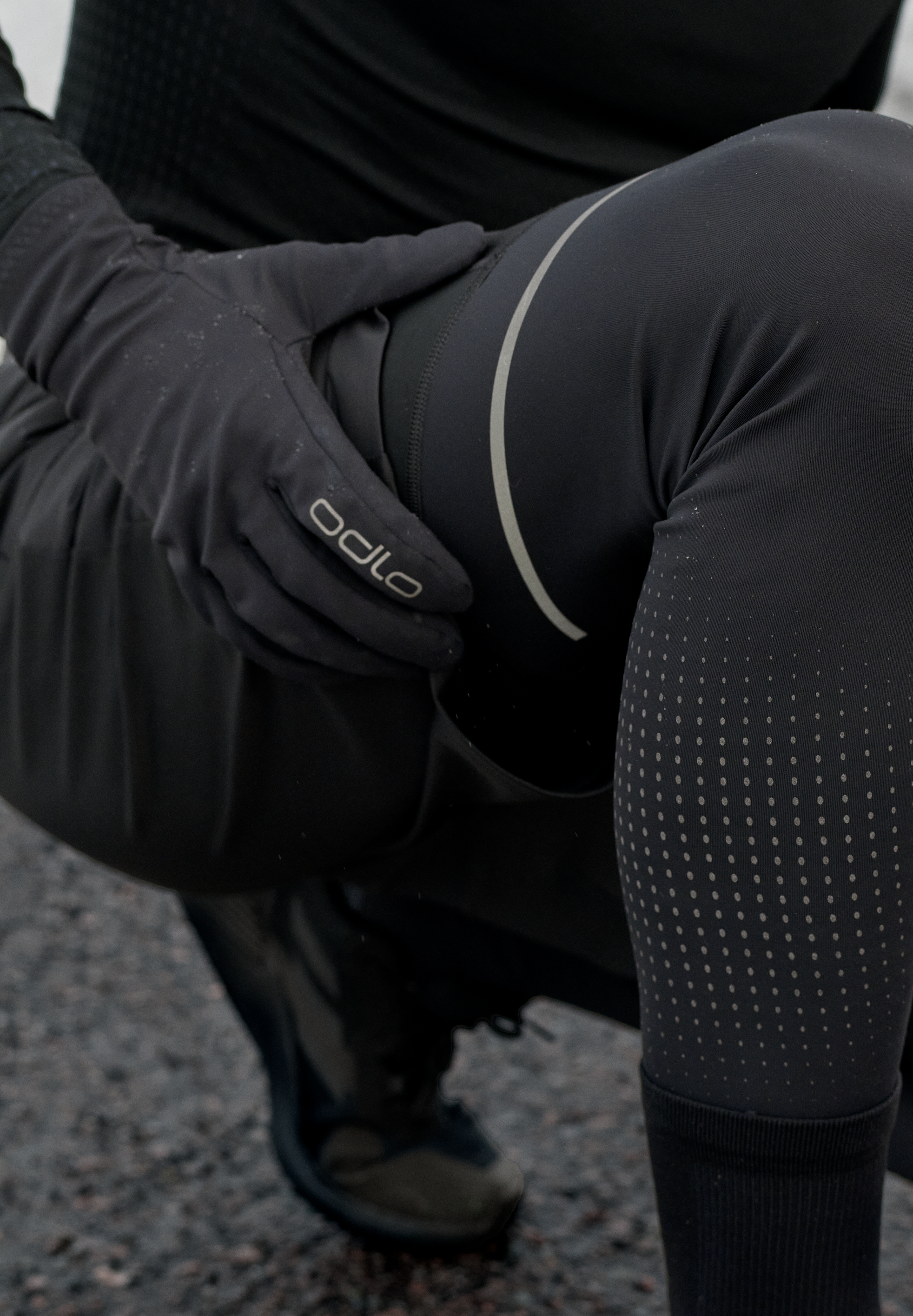 Gehurkt persoon in zwarte Odlo-sportkleding, met handschoenen en legging met reflecterende stippen en een gebogen streep aan de zijkant.