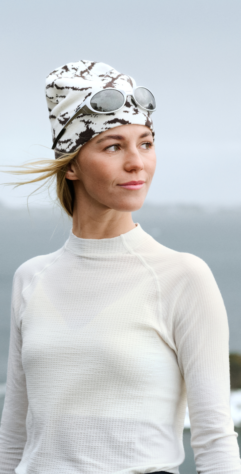 Woman in white top and beanie with sunglasses looks right. Blurry person in foreground. Overcast sea background.