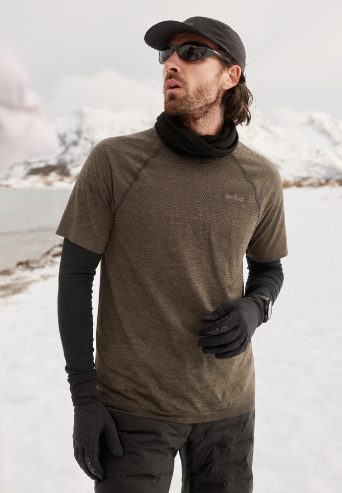 Man in snow wearing a brown Odlo t-shirt over black long sleeves, black gloves, neck gaiter, cap, and sunglasses.
