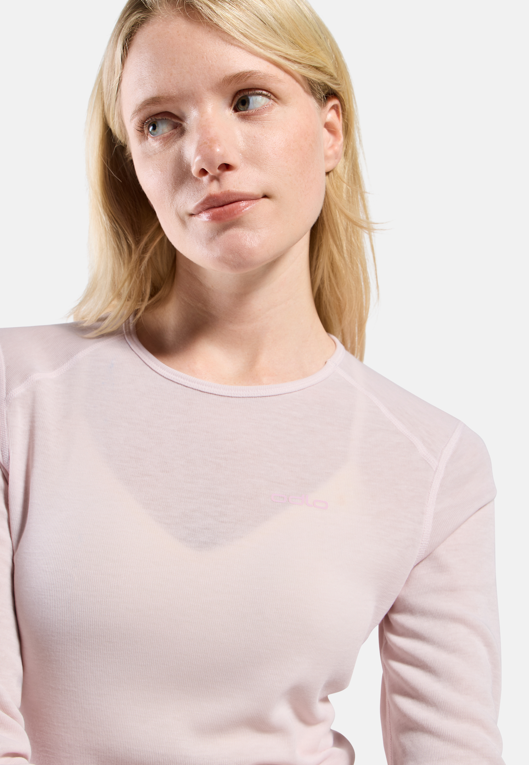 Close-up of a woman looking to the side, wearing a light pink long-sleeve base layer top with subtle logo, against a plain light background.