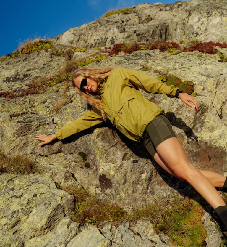 Blonde woman in sunglasses, yellow jacket and green shorts leans back against a rocky, mossy hillside.