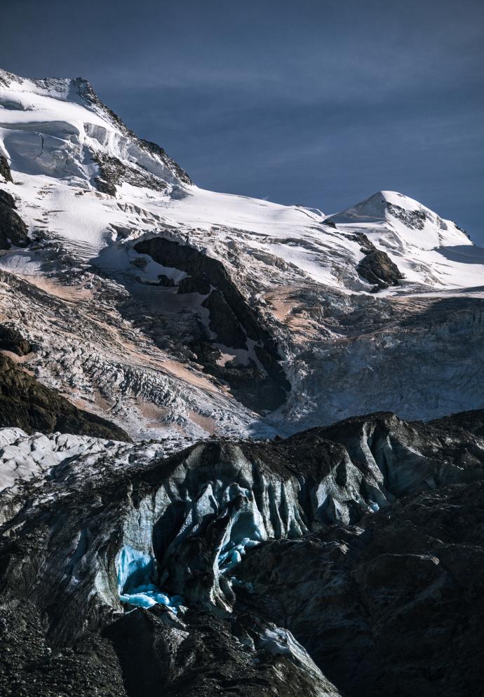 Besneeuwde bergen en donkere rotshellingen boven een ruig gletsjerlandschap, met diepblauwe ijsspleten onder een donkere lucht.