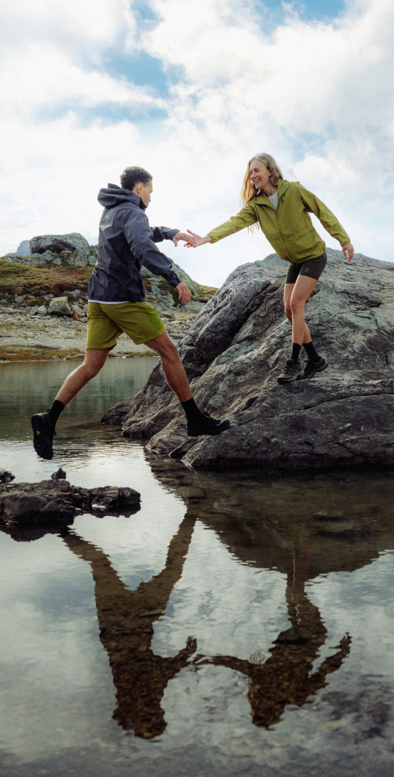 A man in a black jacket jumps across a mountain lake to a rock, where a woman in green reaches out to help.