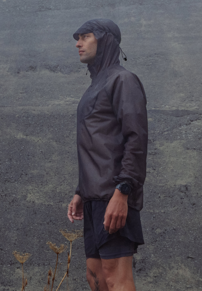 Mann in einer dunklen Kapuzenjacke und kurzen Hose steht im Profil vor einer nebligen, grauen Felslandschaft mit vertrockneten Pflanzen.