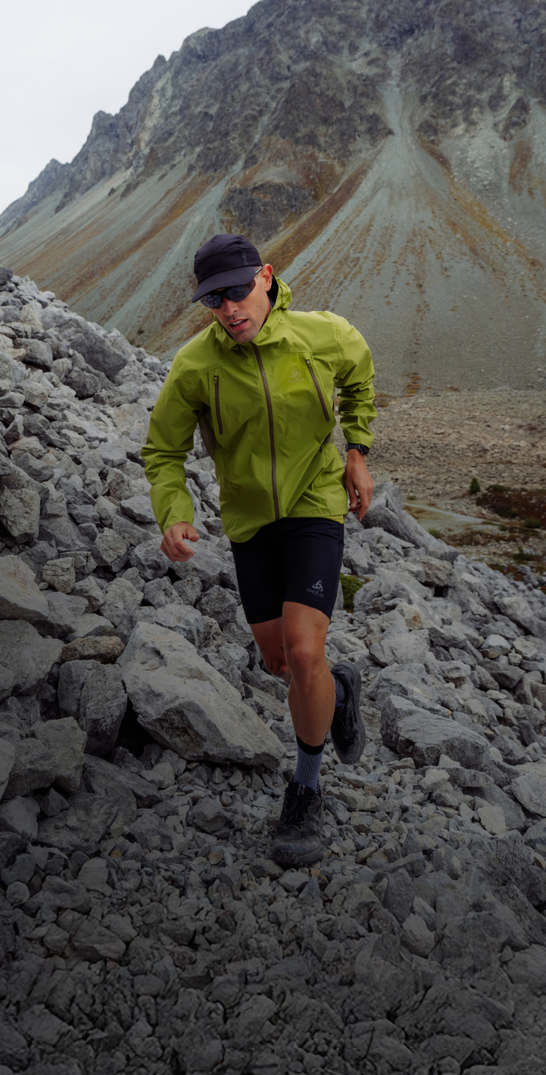 A man in a green jacket, black shorts, cap and sunglasses runs down a steep, rocky mountain slope.