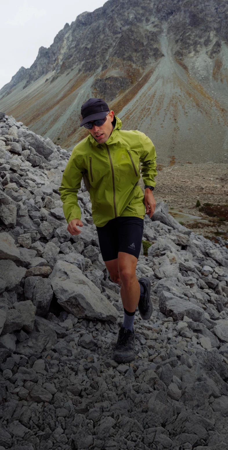 A man in a green jacket, black shorts, cap and sunglasses runs down a steep, rocky mountain slope.
