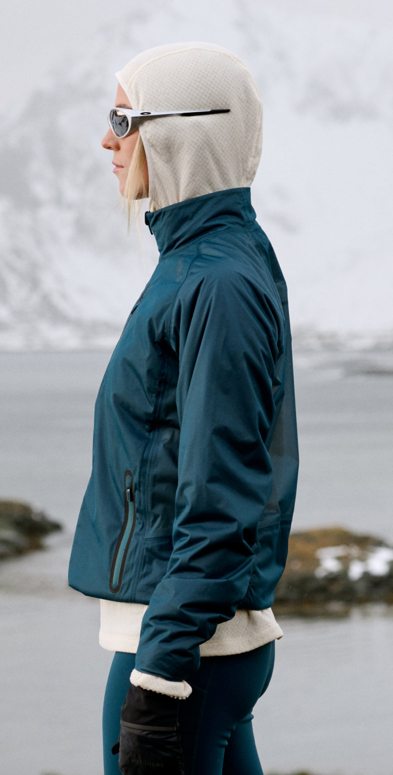 Woman in profile wearing a teal jacket, white hood and sunglasses, standing by water with snowy mountains behind.