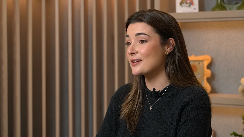 Person with long dark hair speaking indoors, wearing a black top, seated against a softly lit background with shelves