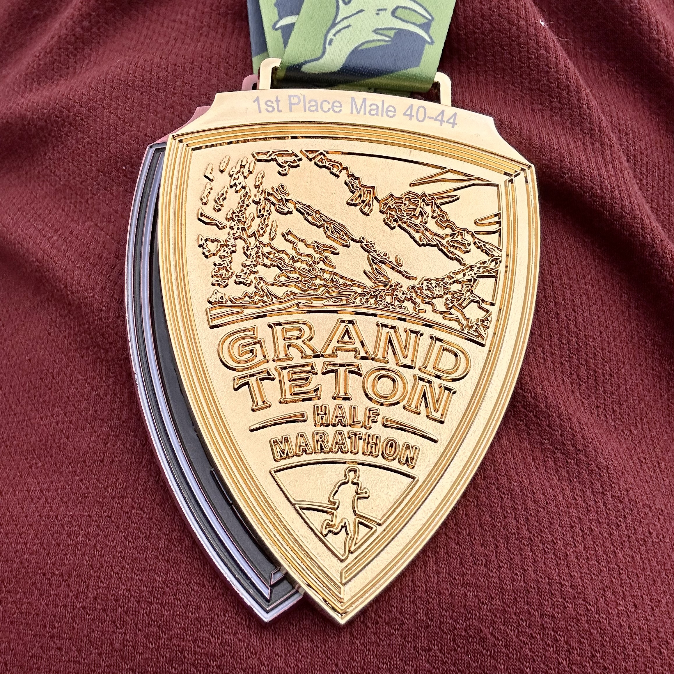 A close-up of the gold medal for the Grand Teton Half Marathon. The medal has "1st place male 40-44" printed at the top, and is embossed with an image of Cascade Canyon, the words "Grand Teton Half Marathon," and a running person.
